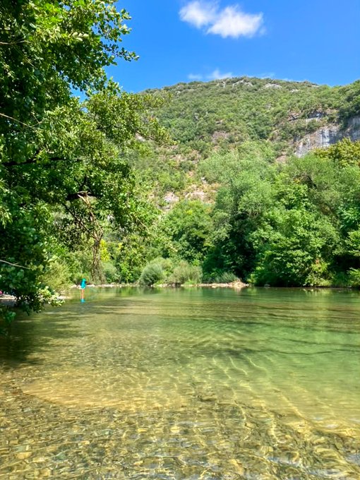 A serene photograph of a clear turquoise river with green mountains and trees under a bright blue sky.