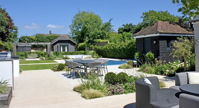 A beautifully landscaped backyard garden featuring a stone patio with outdoor dining furniture, surrounded by lush plants, flowers, and garden sheds.