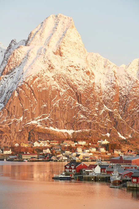 A scenic view of a small fishing village with red rorbuer cabins nestled at the base of a massive snow-covered mountain, reflected in calm fjord waters bathed in warm golden light.