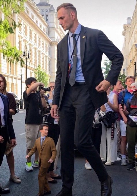 A tall man in a dark suit walks down a European street alongside a notably short man in a brown suit, both styled in formal business attire. The "Not edited btw" caption highlights the striking height difference between the two subjects, emphasizing that the dramatic contrast in their stature is real and unaltered.