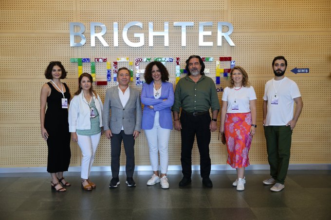 A group of seven people posing for a photo in front of a wooden wall with the word 'BRIGHTER' displayed in large white letters above a mosaic artwork.