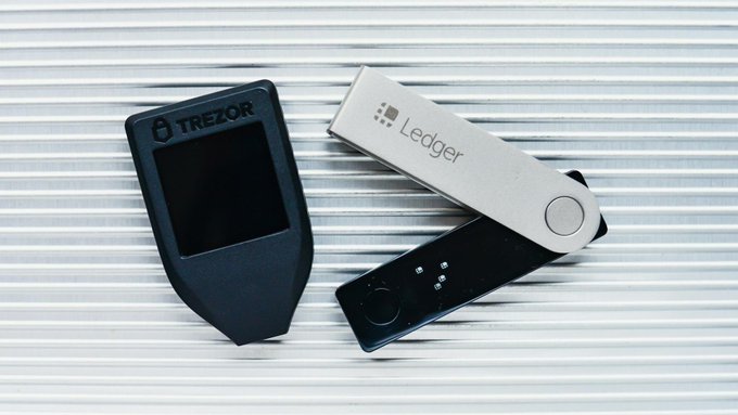 Two cryptocurrency hardware wallets, a black Trezor and a silver Ledger, arranged on a silver corrugated surface.