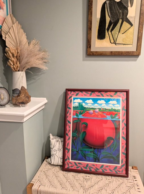 A framed artwork featuring a red tomato and a colorful landscape sits on a woven stool, displayed next to a vase of dried flowers and under a framed animal artwork.