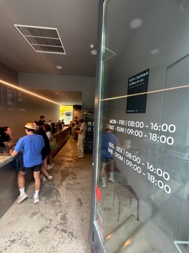 An indoor restaurant or food court area with people queuing, featuring operating hours posted on a glass window: MON-FRI 09:00-16:00 / 17:30-18:00 and SAT-SUN 09:00-16:00.