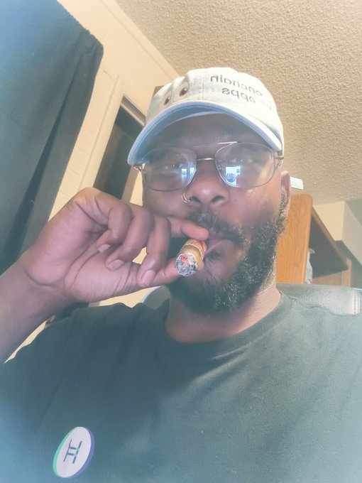 The image features a man wearing glasses, a hat, and a black shirt who is holding a cigar up to his mouth while taking a selfie with the caption "Apecoin A Supporting #Day544 @apecoin Blessings ApE". The man's attire suggests that he might be attending an event or gathering related to the #Day544 hashtag.