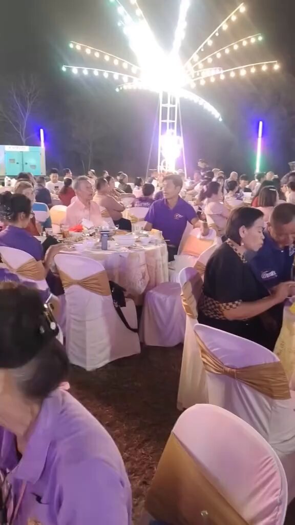 The image shows a large group of people gathered around dining tables at an outdoor event with white chairs and purple tablecloths. The crowd appears to be enjoying themselves, possibly attending a festival or celebration. There are several people sitting at the tables, engaged in conversation and eating food. A few individuals can also be seen standing near the tables.

The scene is set against a backdrop of a large fireworks display, creating an exciting atmosphere for the event. The image captures the essence of a festive gathering where people come together to enjoy each other's company and share a meal under the captivating night sky.