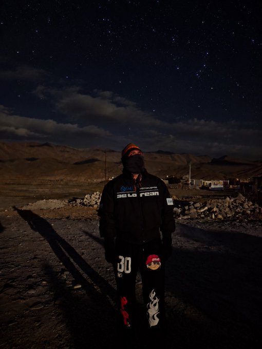 A nighttime photograph of a person standing in a barren, rocky landscape under a starry sky, wearing a beanie, dark jacket with 'Belo Team' text, and black pants featuring flame designs and the number '30'.