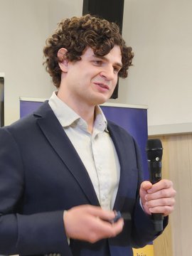 The image features a man wearing a suit jacket and holding a microphone. He appears to be speaking or presenting something, possibly related to finance or investing. The background of the image shows a white wall with a poster hanging behind him.
