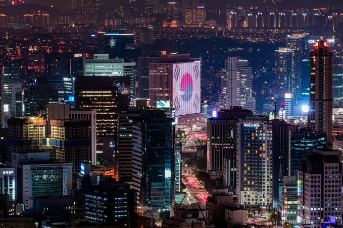 A nighttime aerial view of a bustling Seoul cityscape, featuring illuminated skyscrapers with visible signage for "GLOBAL EDU" and "DFW" alongside a digital billboard, reflecting the vibrant urban financial district. The image visually anchors the post's report on South Korea's crypto trading surge amid a sharp KOSPI decline, illustrating the economic backdrop where investors shifted from equities into digital assets like USDT.
