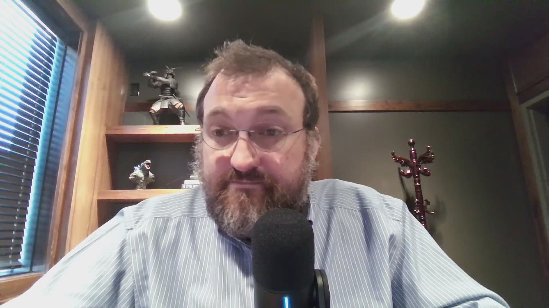 The image shows a man wearing glasses sitting at a desk, holding a microphone and looking into the camera. The man appears to be a speaker or host of a podcast, as he has a microphone in front of him. The background features a window with blinds, suggesting that it might be daytime. There is also an object on the wall behind the man, which could be related to his podcasting setup.