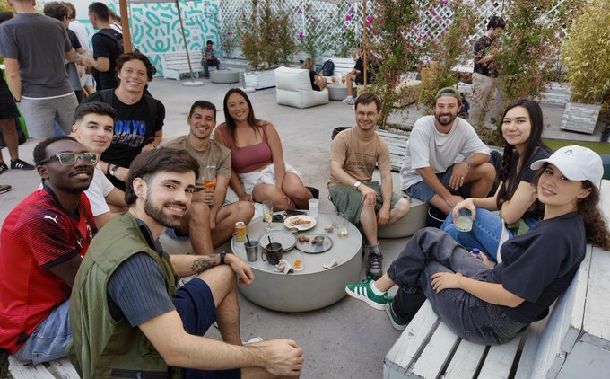 A group of around ten friends sit together on outdoor lounge furniture around a low table, posing for a photo at what appears to be a social gathering.