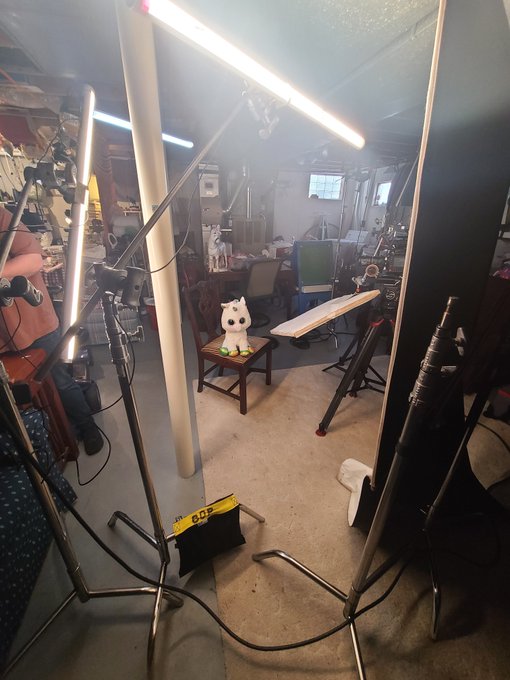 A basement or workshop photo studio setup featuring two long LED light tubes on stands, with a small white dog figurine sitting on a table in the center.