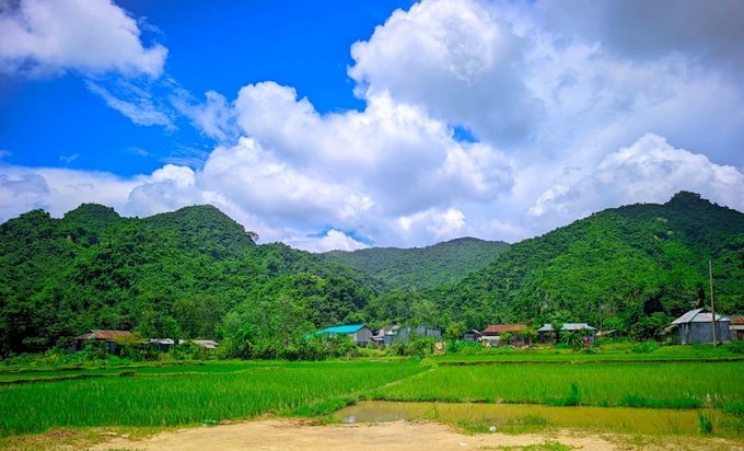 A vibrant rural landscape featuring lush green rice paddies in the foreground, a small village nestled at the base of forested karst mountains, and a dramatic blue sky filled with billowing white cumulus clouds.