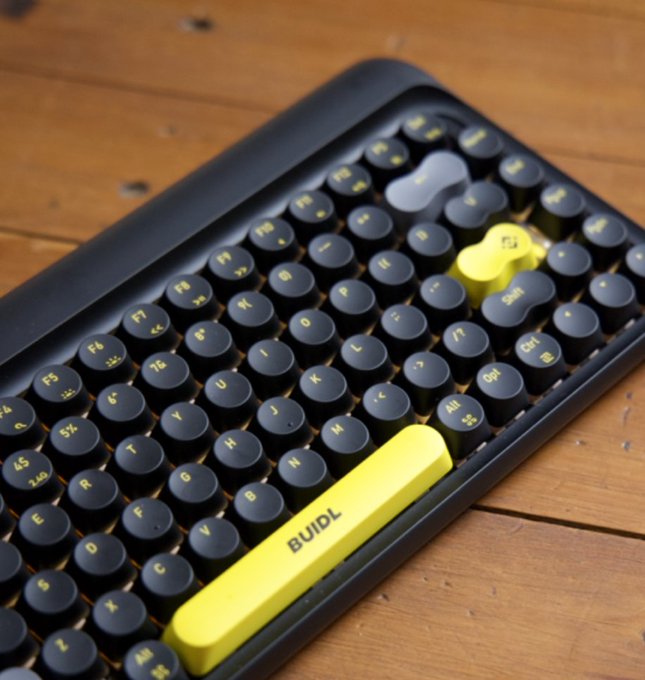 A close-up of a black mechanical keyboard with round keycaps resting on a wooden surface, featuring a yellow spacebar with "BUIDL" printed on it.
