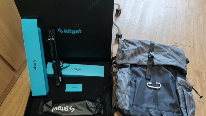 A Bitget branded setup is displayed on a black surface, including a turquoise long box, a smaller turquoise box, a black tube, a black pouch, and a gray checkered backpack, all next to wooden flooring.