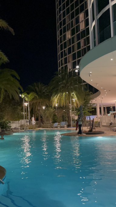 A nighttime view of an illuminated resort swimming pool surrounded by palm trees and a tall building.