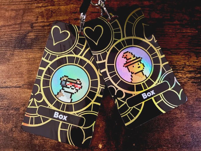 Two holographic keychain tags labeled 'Box' featuring pixelated character portraits within golden circular designs, displayed on a wooden surface.