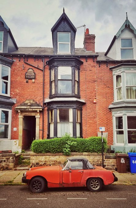 A vintage red convertible sports car is parked on a street in front of a row of traditional red brick Victorian-style houses, with a blue recycling bin visible on the sidewalk.