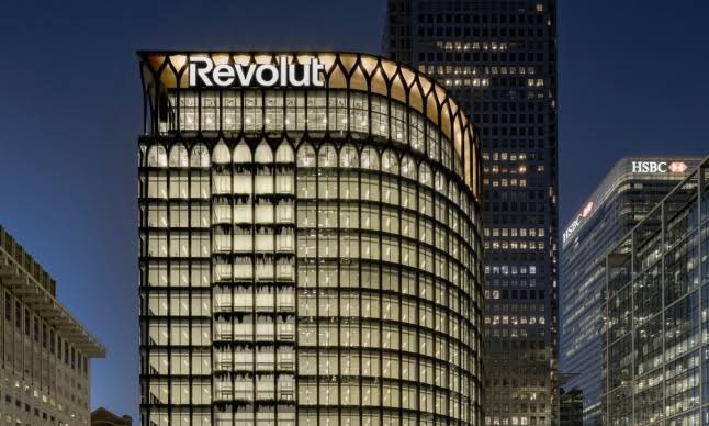 A nighttime photograph of the illuminated Revolut building in London's financial district, with HSBC visible in the background.