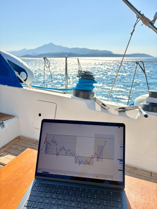 A laptop displaying market charts is set up on the cockpit table of a sailboat, with sparkling blue water and distant mountains creating a serene backdrop for "GM Crew!" trading from the sea.