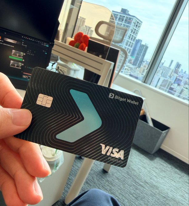 A hand holds up a sleek black "Bitget Wallet" Visa Platinum card in a high-rise office with city views, illustrating the post's point about using the crypto-linked card conveniently in Japan to spend USDT directly without constant yen conversions.