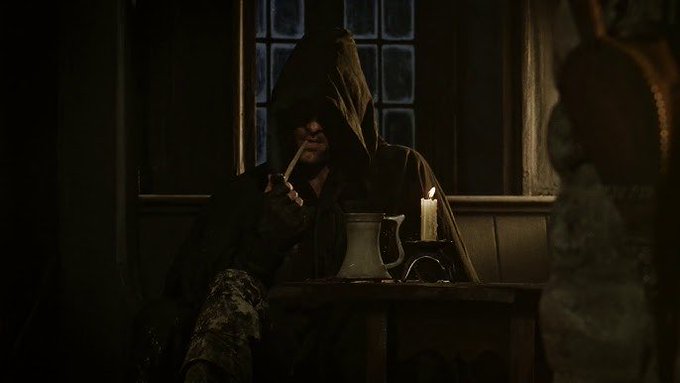 A hooded figure sits at a dimly lit wooden table with a single candle, a pitcher, and a mug, wearing gloves and holding a cigarette.