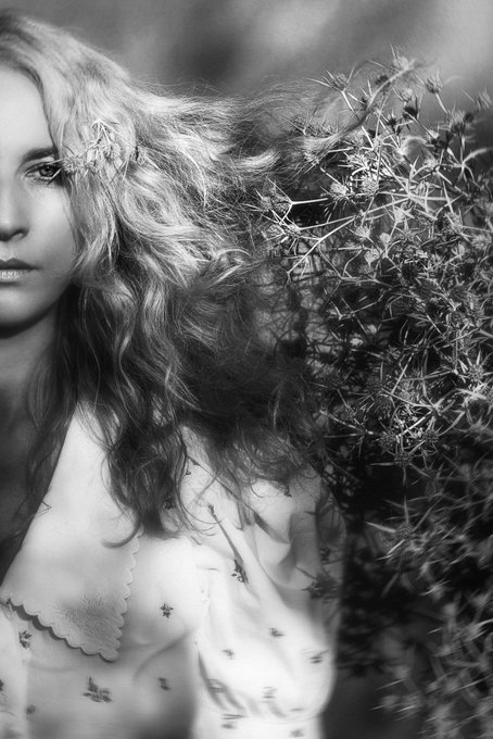 A black and white portrait of a woman with long, curly blonde hair blowing in the wind, wearing a light-colored patterned blouse, with foliage visible in the background.