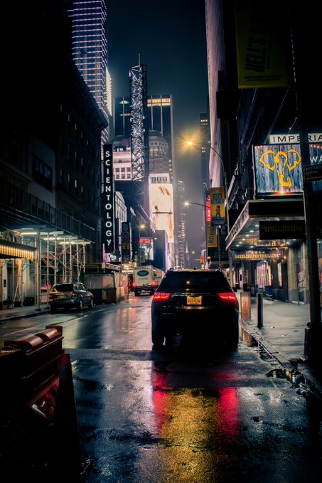 A nighttime city street scene in what appears to be Times Square, showing a car driving down a wet road with illuminated billboards, a Scientology sign, and theater marquees including one for 'SPEAKEASY MAGIC'.