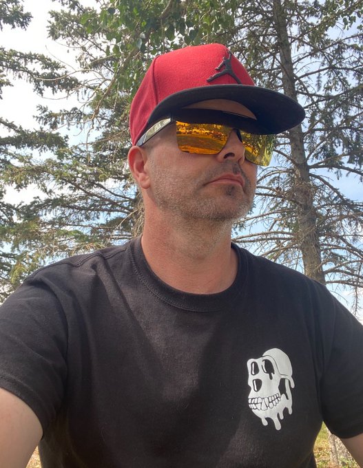 A selfie of a man outdoors wearing a red cap, yellow reflective sunglasses, and a black t-shirt featuring a CryptoPunk-style skull dripping graphic.