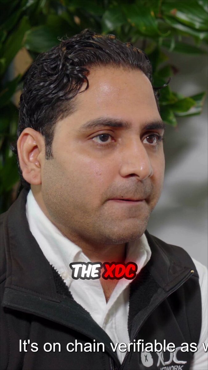 A man wearing a black jacket is standing next to a plant, inside an office setting with other offices visible in the background. The man appears to be talking about the XDC Network or blockchain technology. The post emphasizes that the network helps real-world finance by revolutionizing trade, which is highlighted as a "Must-watch for tech enthusiasts". This description provides context and relevance to the image of the man wearing a jacket next to a plant in an office.