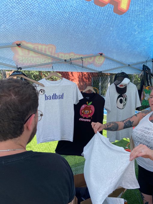 An outdoor vendor booth under a blue tent displaying t-shirts with designs including the word 'badbsd' and cartoon characters, while a person holds up a gray shirt.