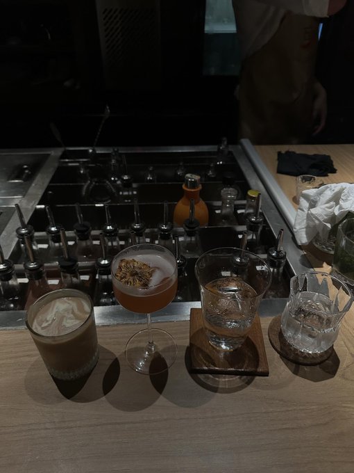 Four cocktails in various glassware sit on a wooden bar counter in front of a metal caddy holding bottles and pour spouts, with a bartender partially visible in the dark background.