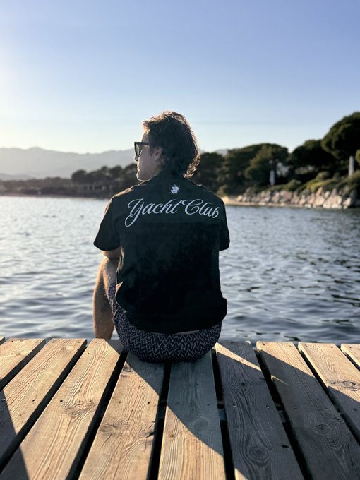 A person sits on a wooden dock overlooking a calm body of water, wearing a black t-shirt with 'Yacht Club' written in cursive on the back.