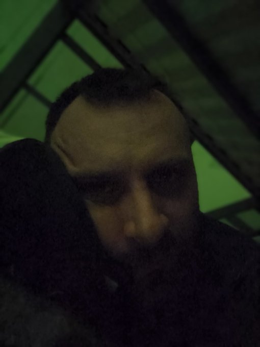 A dark, moody selfie of a person with a beard wearing a hoodie, photographed against a green-lit background.