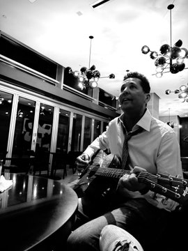 A young man in a dress shirt and tie plays an acoustic guitar while seated at a table, captured in black and white in what appears to be an upscale venue. The image reflects the post's "Work | Hustle | Win" message by portraying a moment of creative passion and focus that complements a driven, ambitious lifestyle.
