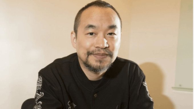 A man with a goatee wearing a black shirt poses for a portrait against a plain wall.