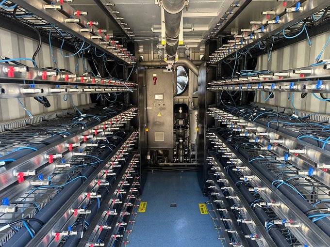 A photograph of an industrial facility interior with rows of pipe racks, cables, and valves flanking a central walkway.