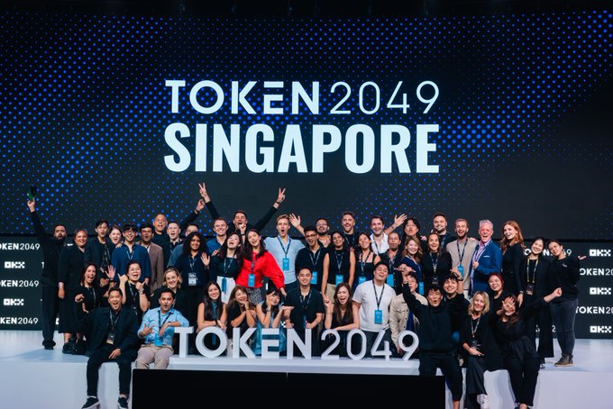 A large group photo of conference attendees on stage at 'TOKEN 2049 SINGAPORE', with participants smiling, making hand gestures, and posing in front of a large branded backdrop.