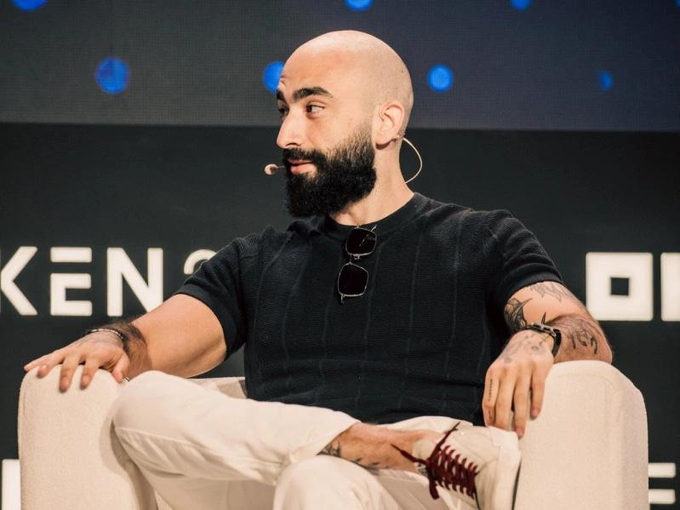 A bald, bearded man wearing a black sweater and white pants sits on a chair on a stage, with a partial 'KEN' logo visible in the background.