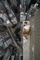 A person in an orange dress poses on a narrow ledge between tall buildings, with auction bids of 0.5 ETH noted for the Horizon & Liminal artworks by Angela Nikolau. The image captures the artist's signature rooftop photography style, reflecting the daring nature of her work.