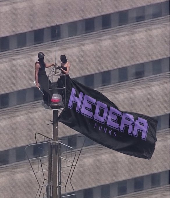 Two people in black stand on a high antenna or tower platform, unfurling a large black banner reading 'HEGE... PUNKS' in purple text against a building facade.