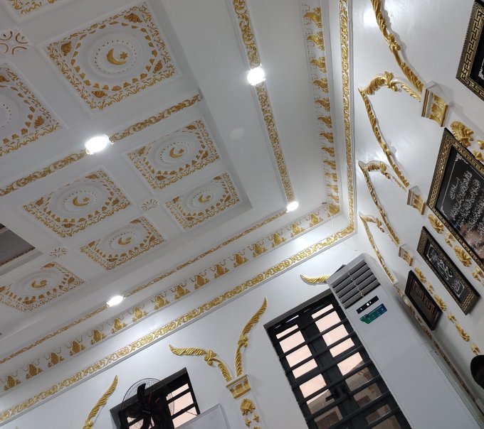 An ornately decorated ceiling with white and gold geometric patterns and crescent moon motifs, alongside framed Islamic calligraphy on the wall.