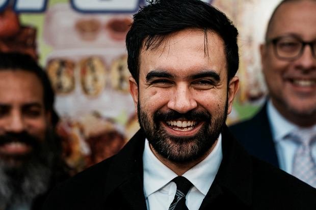 A smiling man with dark hair and a beard wearing a black coat and tie, photographed at what appears to be an event, with two other men visible in the background.