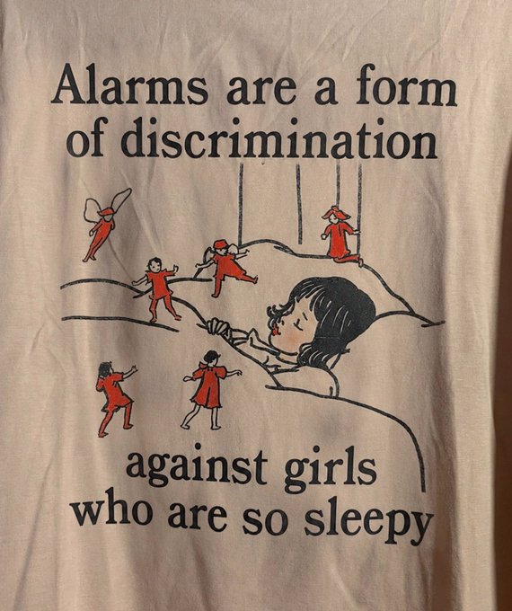 Cream-colored t-shirt with vintage-style text reading 'Alarms are a form of discrimination against girls who are so sleepy', showing a sleeping girl with tiny red-clad figures dancing on her pillow.