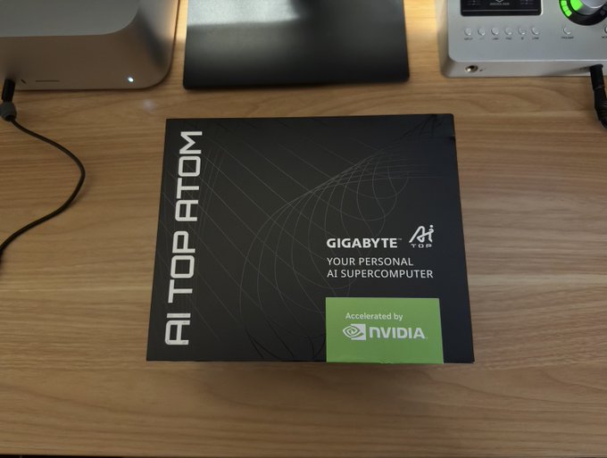 A black product box for a 'GIGABYTE AI TOP ATOM' personal AI supercomputer, noting it is 'Accelerated by NVIDIA', sits on a wooden desk next to other electronics.