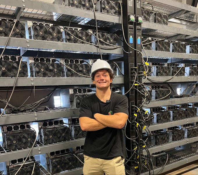 A smiling young man wearing a white hard hat and black t-shirt stands with arms crossed in front of large metal racks filled with numerous crypto mining rigs featuring cooling fans and tangled power cables.