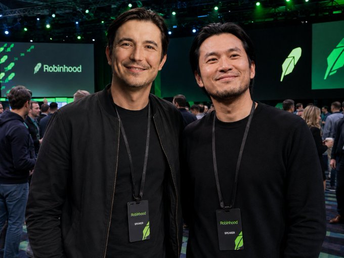 Two men wearing "Robinhood Speaker" lanyards pose together at what appears to be a Robinhood event, with the company's feather logo visible on screens behind them. The image humorously illustrates the post's cautionary tale about trusting friends' crypto tips—since the company running this conference (whose logos are on full display) famously had co-founders who made early bets on Bitcoin and Nvidia that paid off enormously, making the poster's brag about their "friend's" recommendations especially ironic.
