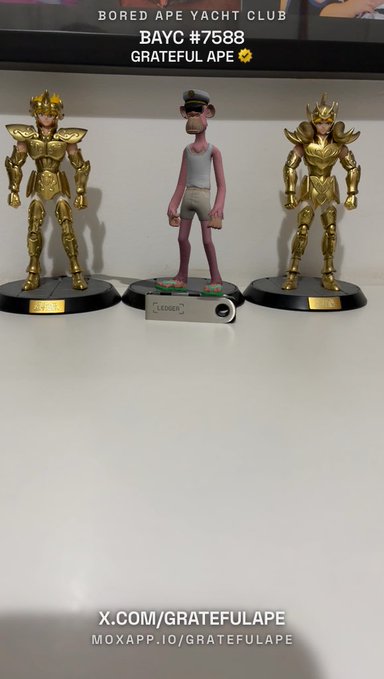 Three figurines displayed on a shelf: two gold-armored warriors flanking a central lanky character with a 'LEADER' tag, labeled 'BORED APE YACHT CLUB BAYC #7588 GRATEFUL APE' with social media handles.