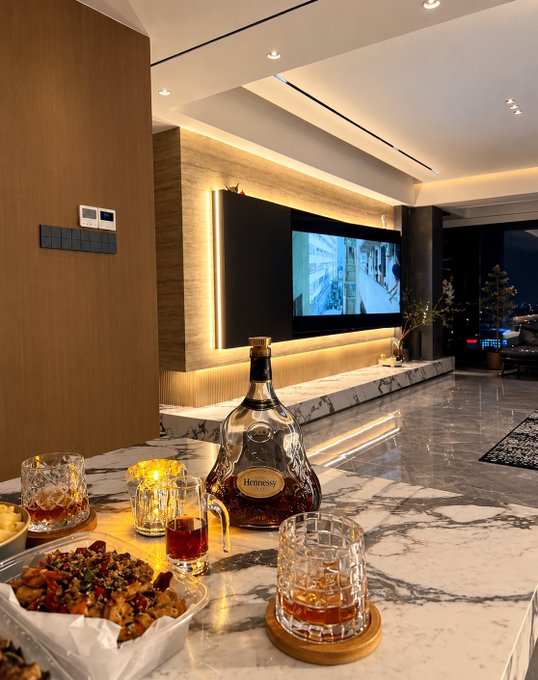 A luxury living room features a marble bar counter displaying a bottle of Hennessy cognac, whiskey glasses, and snacks, with a wall-mounted TV and ambient lighting in the background.