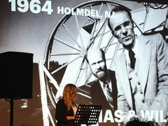 A presenter stands at a podium in front of a projected slide showing a black-and-white photo from 1964 in Holmdel featuring Arno Penzias and Robert Wilson with the Holmdel Horn Antenna, labeled "IASA MIL..."